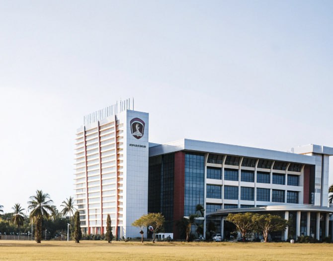 Modern university campus building with a tall tower featuring the institution’s logo, large glass windows, palm trees, and an open lawn in the foreground.