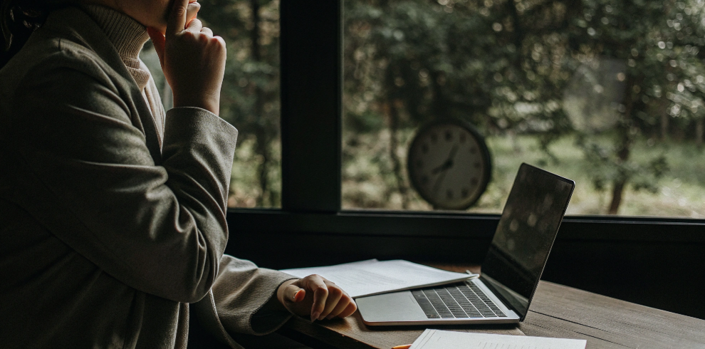 Student sitting at a desk with laptop and documents, thinking about exam results and career planning while looking out the window.